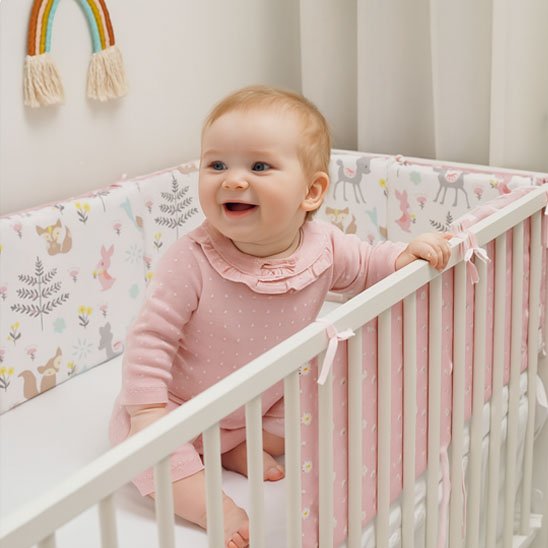Bébé fille en rose debout dans son lit avec tour de lit coton doux motif animaux rose pâle
